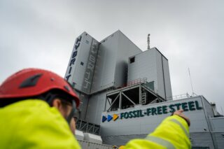 A worker in a hard hat and high visibility coat in the foreground points to a large factory emblazoned with the words "Fossil-Free Steel."