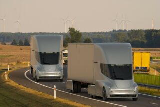 Two large, futuristic-looking electric lorries drive down an open road with wind turbines in the background.